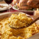 A close-up of a wooden spoon lifting a steaming scoop of creamy, golden corn casserole from a baking dish.