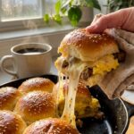 A close-up of a hand pulling a single breakfast slider from the pan, showing a gooey cheese stretch, fluffy egg, and sausage filling.