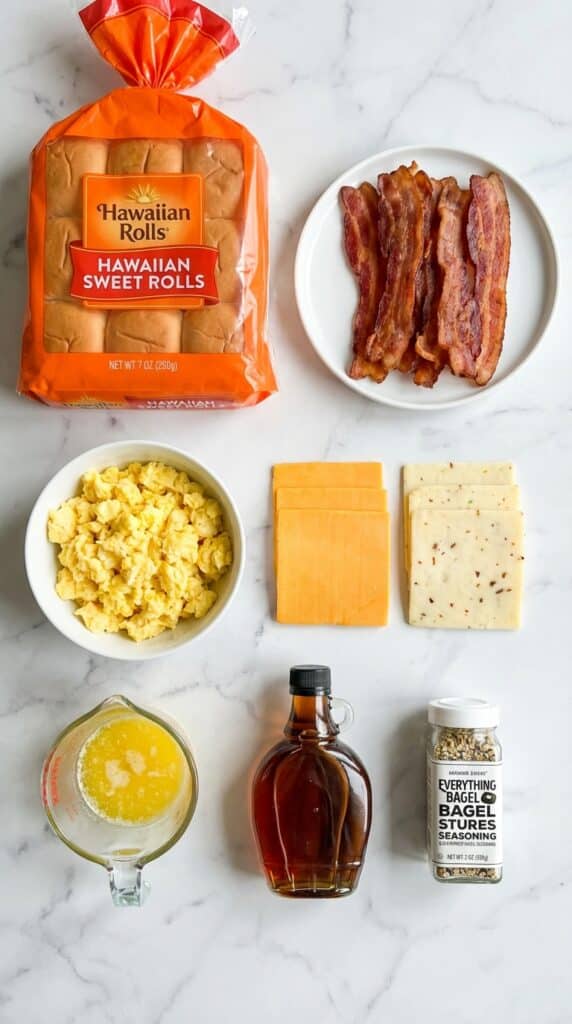 A flat lay showing a pack of Hawaiian rolls, cooked bacon, scrambled eggs, sliced cheese, melted butter, maple syrup, and seasoning on a marble board.