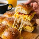 A close-up of a hand pulling a single bacon, egg, and cheese slider from a batch, creating a long, gooey cheese pull.