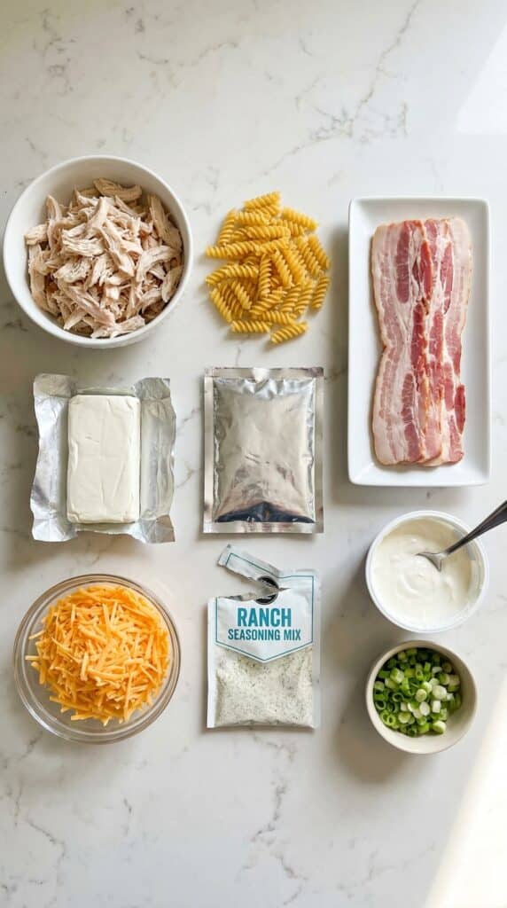 A flat lay view showing ingredients for the casserole: shredded chicken, uncooked pasta, raw bacon, cream cheese, cheddar cheese, ranch seasoning, sour cream, and green onions on a marble counter.