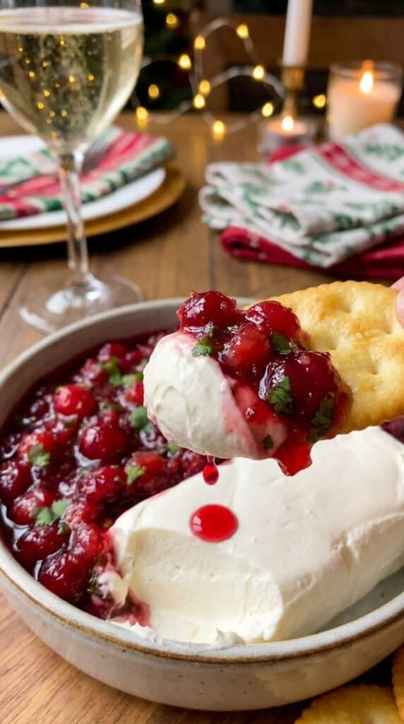A close-up of a buttery cracker scooping up a layer of white cream cheese and glossy red cranberry salsa.