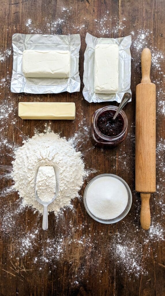 A flat lay showing cream cheese, butter, flour, sugar, a jar of red raspberry jam, and a rolling pin on a floured wooden board.