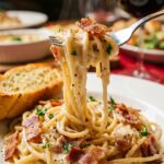 A close-up of a fork twirling penne pasta coated in creamy sauce and bacon bits, with steam rising.