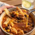 A close-up of a spoon lifting a hearty bite of creamy chicken enchilada soup with a crispy tortilla strip and melted cheese.