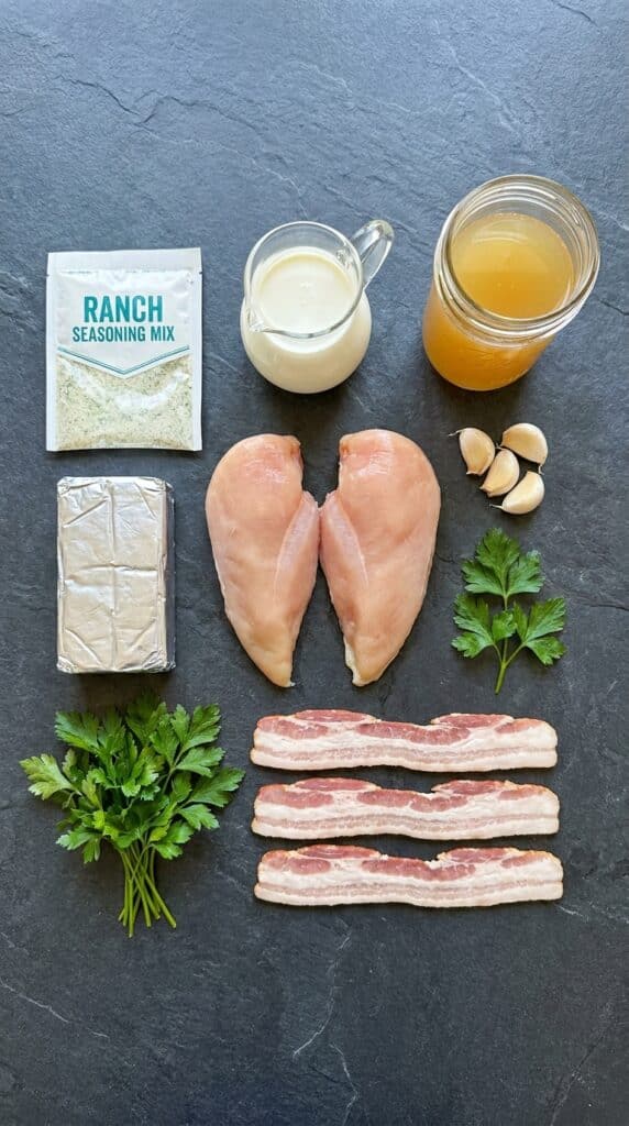 A flat lay showing raw chicken, cream cheese, heavy cream, dry ranch seasoning, chicken broth, garlic, and bacon on a dark slate board.