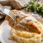 A close-up of a fork cutting through a tender, sauce-covered piece of creamy ranch chicken served over fluffy mashed potatoes.