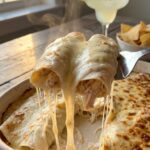 A close-up of a spatula lifting a creamy white chicken enchilada from a baking dish, creating a gooey cheese pull.