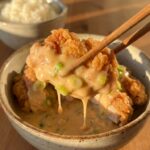 A close-up of wooden chopsticks holding a piece of crispy fried chicken coated in creamy orange sauce, with white rice in the background.