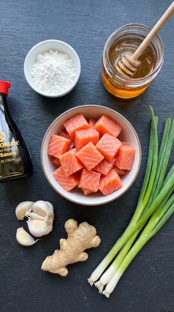 A flat lay showing raw cubed salmon, cornstarch, honey, soy sauce, garlic, ginger, and green onions on a dark slate board.
