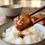 A close-up of wooden chopsticks holding a crispy salmon bite dripping with a glossy honey garlic glaze over a bowl of rice.