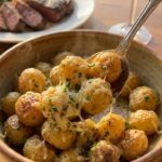 A close-up of a serving spoon lifting buttery, Parmesan-coated baby potatoes from a bowl, with steak in the background.