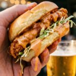 A close-up of a hand holding a messy chicken slider dripping with creamy, spicy orange sauce, with a beer in the blurred background.A close-up of a hand holding a messy chicken slider dripping with creamy, spicy orange sauce, with a beer in the blurred background.