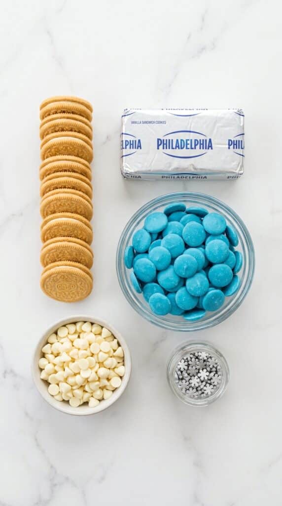 A flat lay showing vanilla cookies, cream cheese, blue candy melts, white chocolate chips, and snowflake sprinkles on a white marble board.
