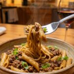A close-up of a fork holding a bite of glossy beef pasta garnished with fresh green onions, with a glass of wine in the background.
