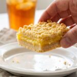 A close-up of a hand holding a lemon crumb bar, showing the layers of shortbread crust, yellow lemon filling, and golden crumble.