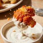 A close-up of a crispy orange buffalo cauliflower bite being dipped into a bowl of creamy ranch dressing.