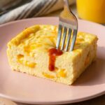A close-up of a fork cutting into a thick, fluffy square of baked eggs with a drop of hot sauce on top.