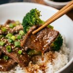 A close-up of wooden chopsticks lifting a piece of glossy beef and broccoli over a bowl of white rice, garnished with fresh green onions.