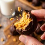 A close-up of a hand holding a chocolate peanut butter ball with a bite taken out, showing the crispy, flaky interior.