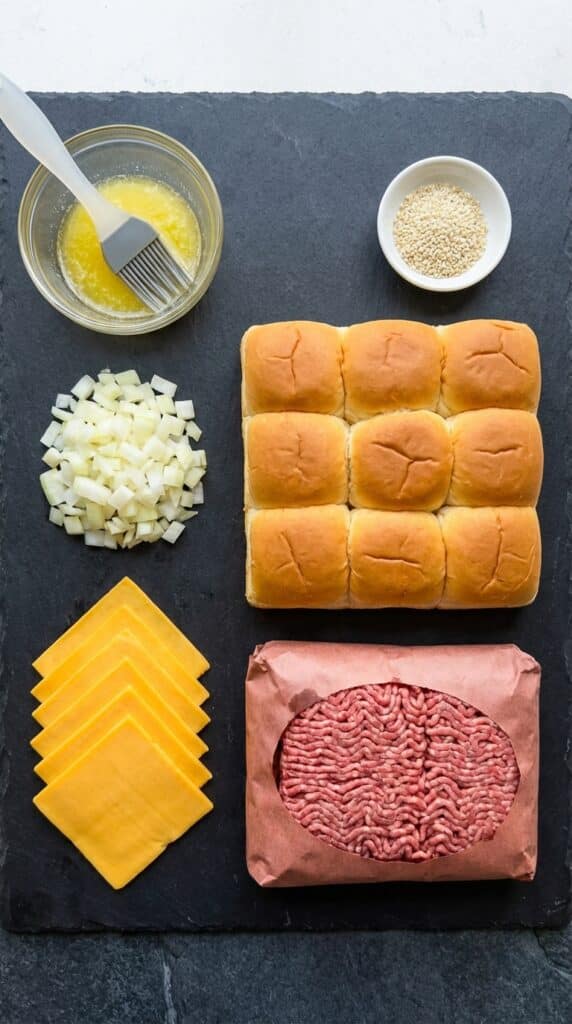 A flat lay showing a connected pack of Hawaiian rolls, ground beef, sliced cheese, onions, and melted butter on a slate board.