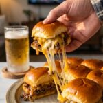 A close-up of a hand pulling a cheeseburger slider from a baked batch, creating a long, gooey cheese stretch.