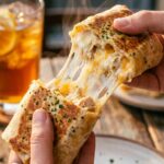 A close-up of hands pulling apart a crispy garlic chicken wrap, creating a gooey cheese stretch from the hot filling.