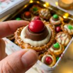 A close-up of a hand holding a single pretzel hug treat, showing the pretzel, melted chocolate, and red M&M layers.