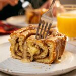 A close-up of a fork cutting into a fluffy square of baked cinnamon roll casserole, revealing the soft interior and the thick glaze dripping down the side.