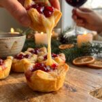A close-up of a hand picking up a cranberry brie bite, creating a stringy cheese pull from the wooden platter.