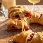 A close-up of a slice of cranberry brie crescent wreath being pulled away, creating a gooey cheese stretch, with festive decor in the background.