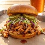 A close-up of a messy pulled BBQ chicken sandwich topped with coleslaw and pickles on a brioche bun.