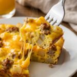 A close-up of a fork cutting into a thick slice of breakfast casserole, showing fluffy eggs, hash browns, sausage, and a gooey cheese pull.