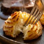 A close-up of a fork cutting into a crispy baked Duchess Potato, showing the fluffy mashed potato interior, with a roast in the background.