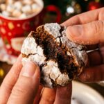 A close-up of a gingerbread crinkle cookie being broken in half to show a soft, chewy center, with a mug of hot chocolate and tree lights in the background.