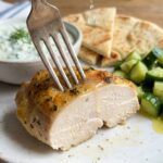 A close-up of a fork piercing a slice of juicy lemon oregano chicken, with lemon juice dripping down, and tzatziki in the background.