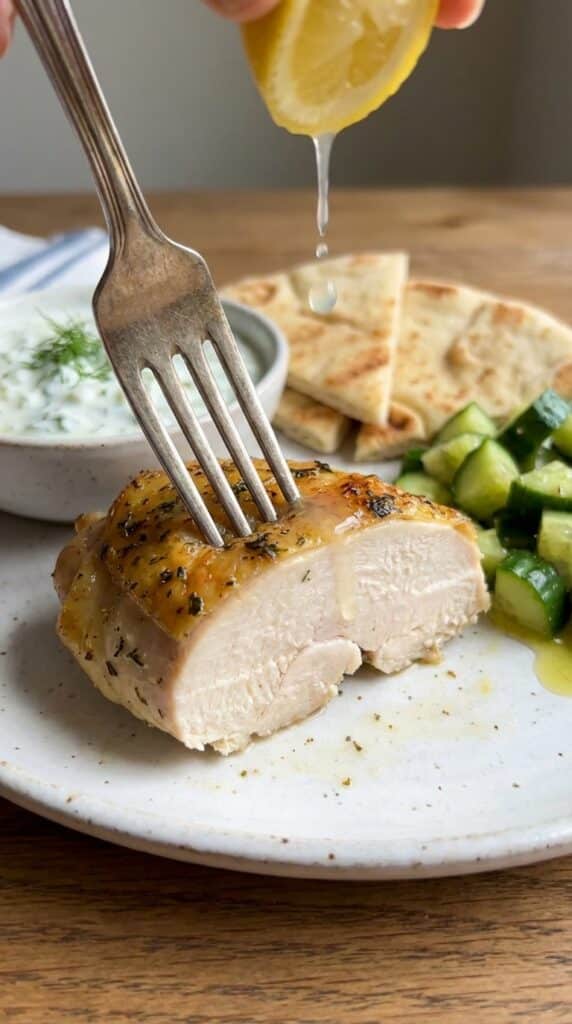 A close-up of a fork piercing a slice of juicy lemon oregano chicken, with lemon juice dripping down, and tzatziki in the background.