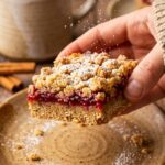 A close-up of a hand lifting a cranberry crumble bar, showing the thick layers of oat crust, red berry filling, and crumble topping, with holiday lights in the background.