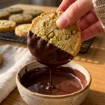 A close-up of a hand dipping a crumbly pistachio shortbread cookie into a bowl of melted dark chocolate.