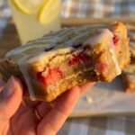 A close-up of a hand holding a dense strawberry lemon blondie with a bite taken out, with a glass of lemonade in the background.