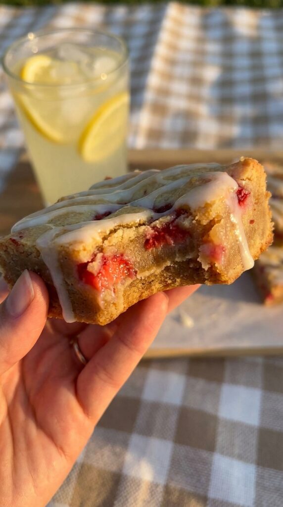 A close-up of a hand holding a dense strawberry lemon blondie with a bite taken out, with a glass of lemonade in the background.
