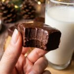A close-up of a hand holding a square of chocolate fudge with a bite taken out, showing its smooth, dense, and creamy interior.
