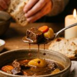 A close-up of a spoon holding a chunk of beef, a carrot, and a potato coated in thick brown gravy, with crusty bread in the background.