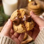 A close-up of hands breaking a warm peanut butter blossom cookie in half, showing a soft center and a slightly melted chocolate kiss.