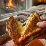 A close-up of a crispy grilled cheese sandwich being dunked into a bowl of hot tomato soup, showing a gooey cheese pull.