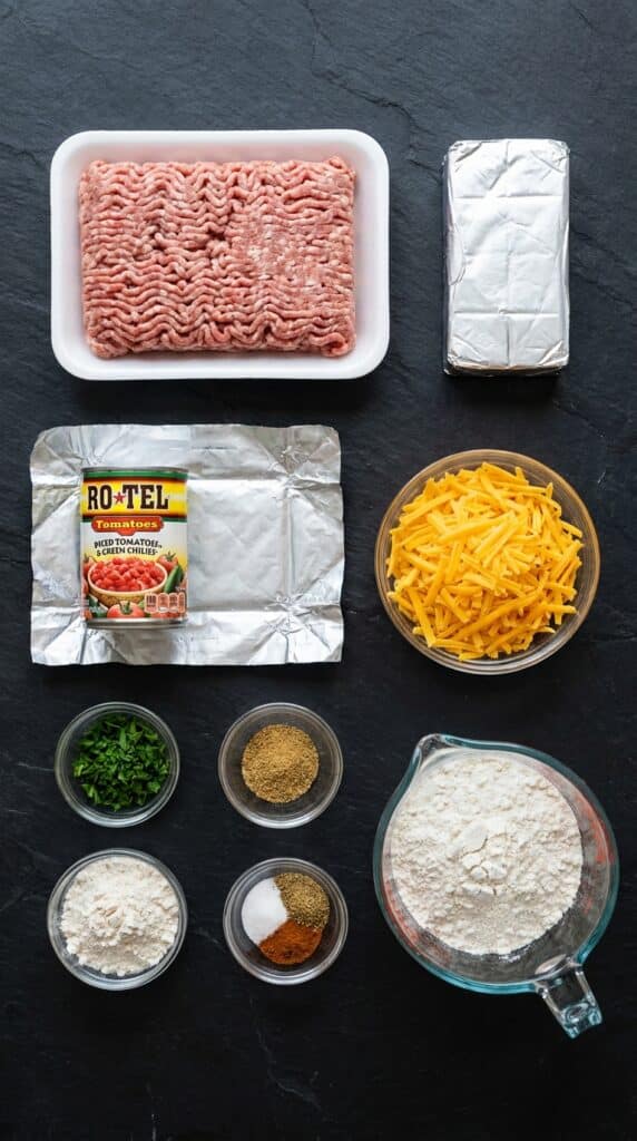 A flat lay showing raw ground sausage, cream cheese, a can of diced tomatoes and green chilies, shredded cheddar, and baking mix on a dark slate board.