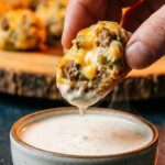 A close-up of a hand dipping a half-eaten, moist sausage ball filled with cheese and green chilies into a bowl of ranch dressing.