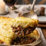A close-up of a serving spoon scooping out a portion of Shepherd's pie, showing distinct layers of mashed potatoes and meat filling.