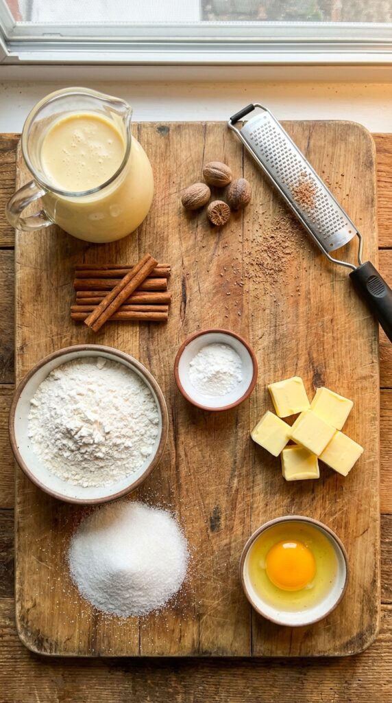 A flat lay showing eggnog, whole nutmeg, cinnamon sticks, butter, flour, sugar, and cream of tartar on a wooden board.