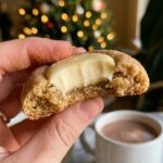 A close-up of a hand holding an eggnog snickerdoodle cookie with a bite taken out, showing the soft center and creamy frosting.